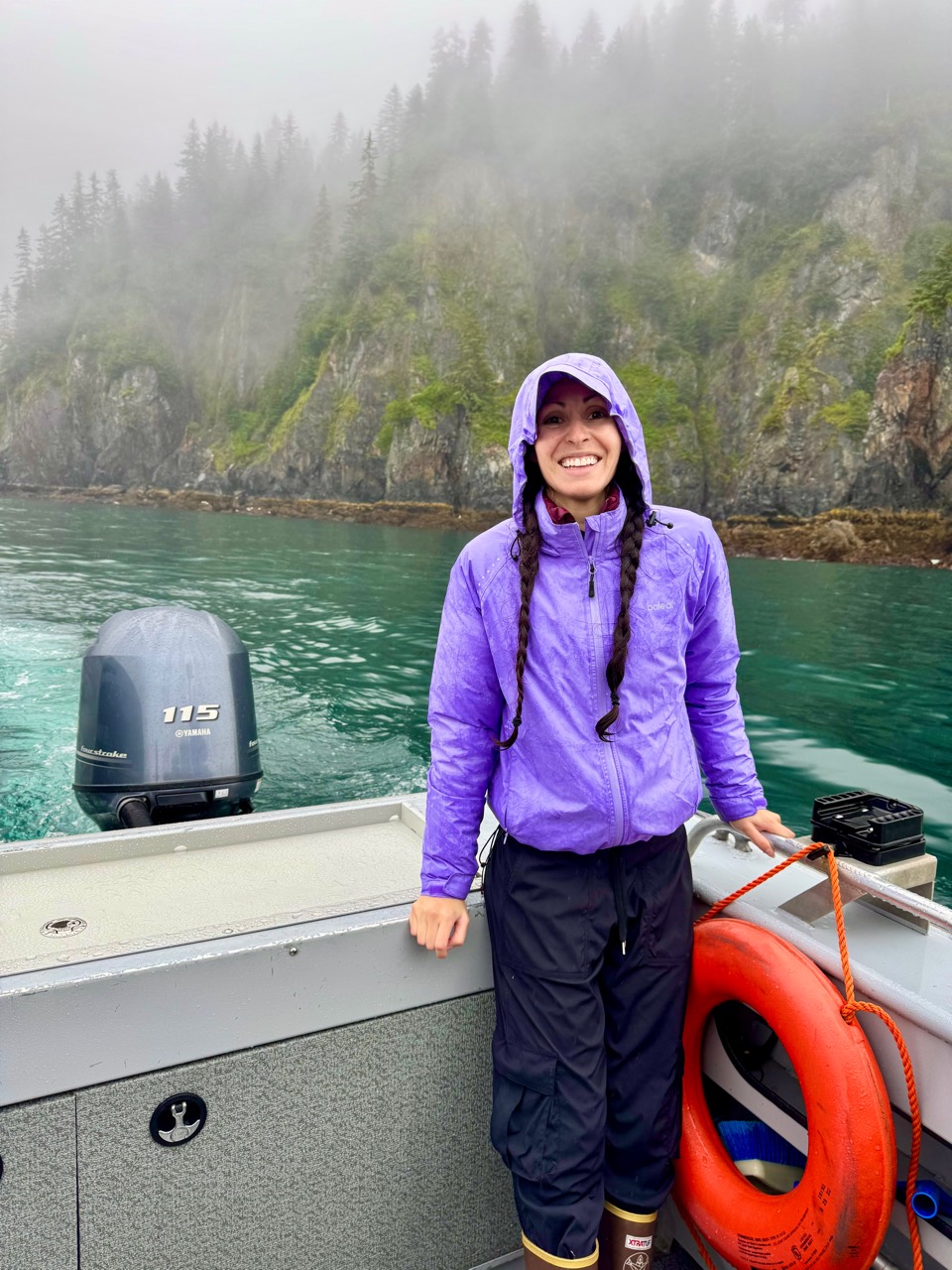 Mona smiling in her purple rain jacket aboard a boat, with misty Alaska cliffs behind her